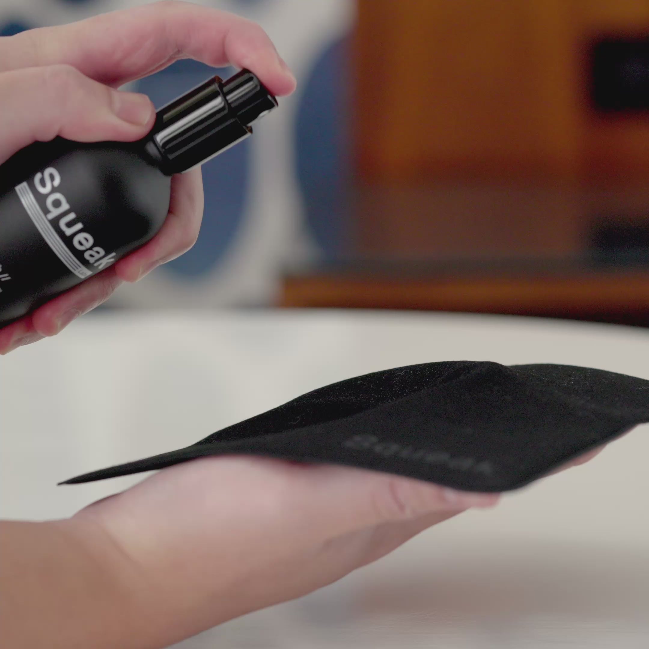 Video of a person spraying Squeak cleaner onto a black Squeak-branded microfiber cloth, preparing to clean a device. The Squeak bottle remains slightly blurred in the background as the cloth is misted.
