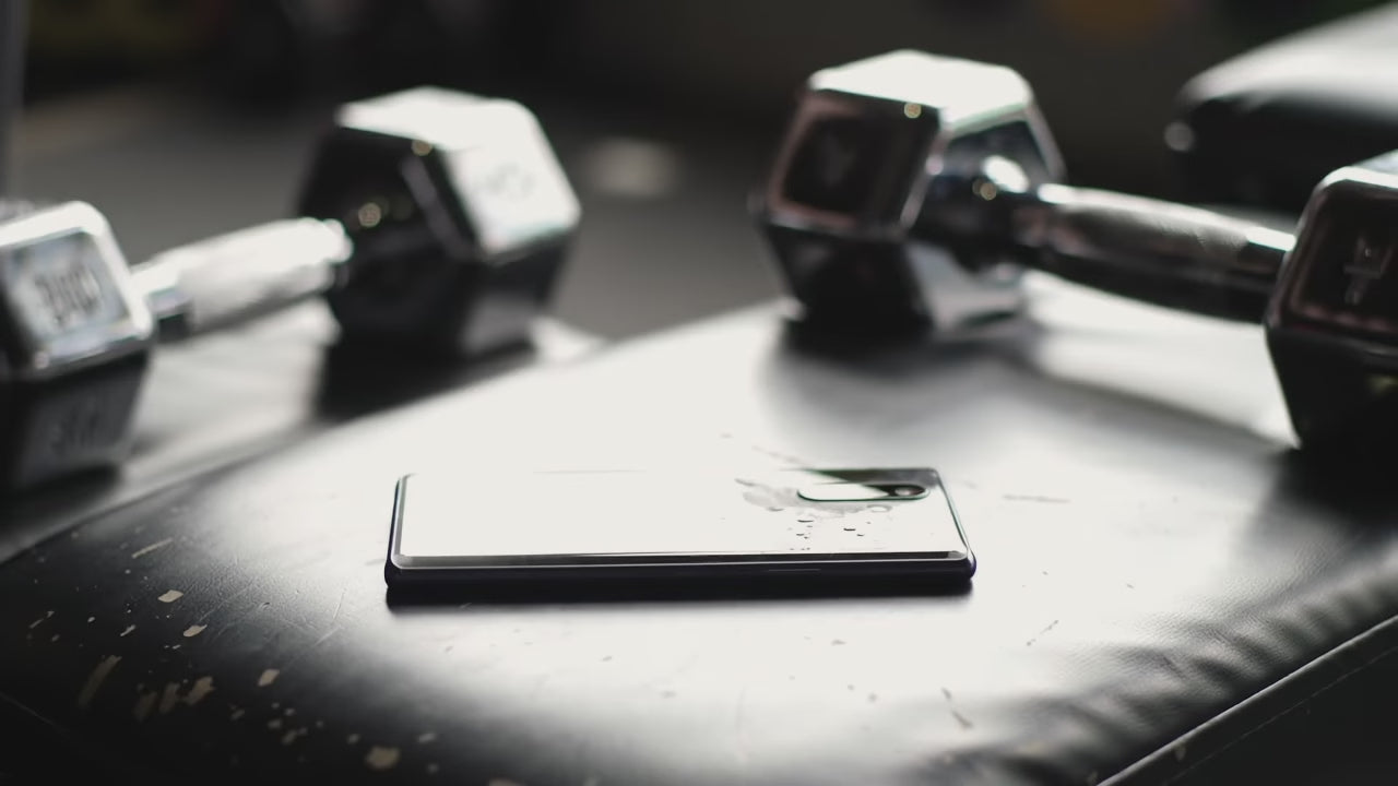 Video: A person picks up a smartphone on a flat gym bench. Dumbbells in the background. Person's sweat drips as they place the phone back down. The person then reaches for the dumbbells.