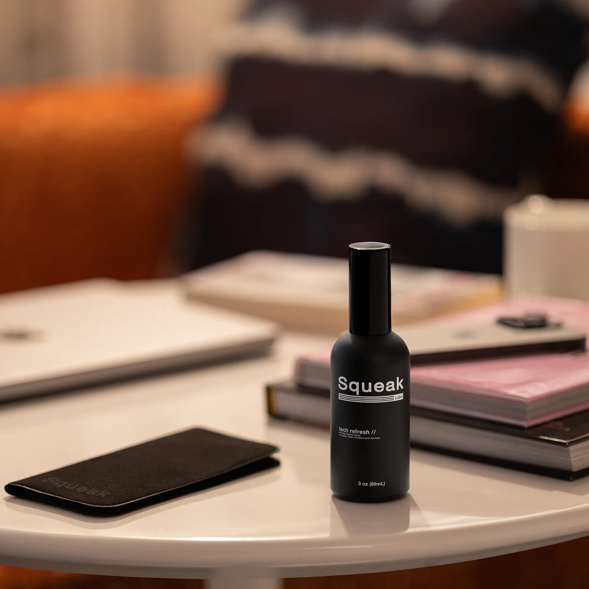 A matte black bottle of Squeak Tech Refresh sits on a white tabletop beside a matching black microfiber cloth. In the softly lit background, a laptop, books, and a mug suggest a cozy, modern workspace.