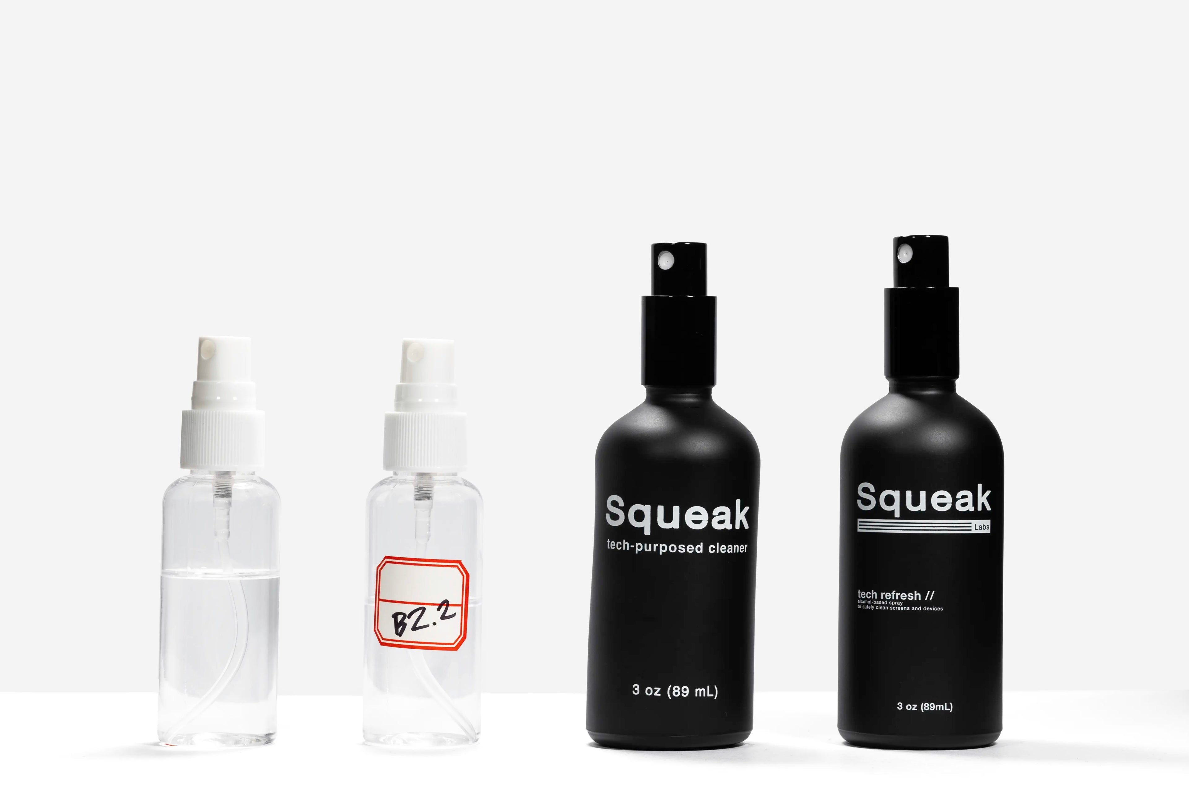 Four spray bottles are lined up to show the product’s development: two small clear sample bottles on the left—one with a handwritten label—and two finished black Squeak bottles on the right, featuring the “tech-purposed cleaner” and “Tech Refresh” 3 oz designs.