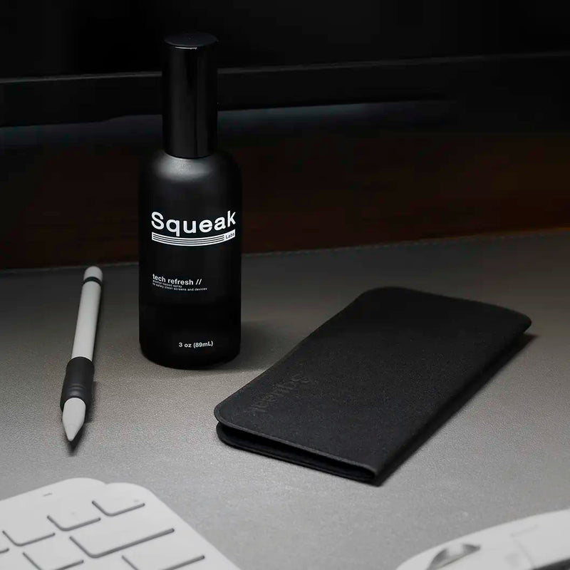 A black Squeak Labs “Tech Refresh” spray bottle sits on a desk beside a folded black microfiber cloth. A stylus, keyboard, and monitor are partially visible, creating a clean workspace setting.