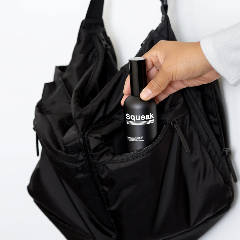A person places a black Squeak Labs “Tech Refresh” spray bottle into the front zipper pocket of a black backpack.