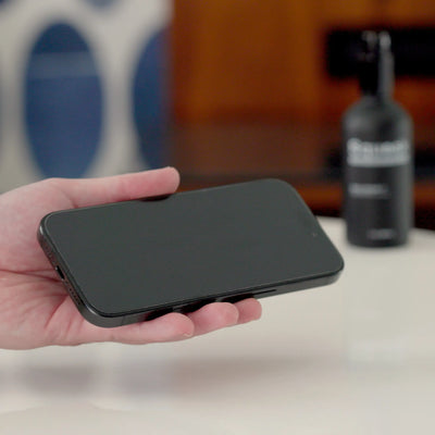 A person holds a shined smartphone with a clean screen in their hand, with a Squeak Labs cleaning spray bottle blurred in the background.